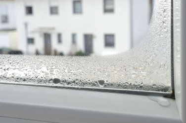 An der unteren linken Ecke einer Glasscheibe in einem weißen Fensterrahmen sammelt sich Kondenswasser in großen Tropfen.