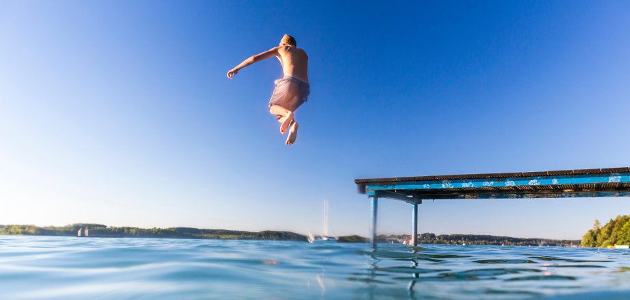 Ein Junge springt bei Sonnenschein von einem Steg in einen Badesee.