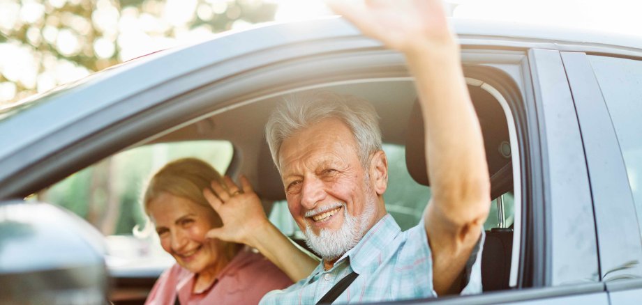 Ein älteres Paar sitzt in seinem Auto und winkt freundlich aus dem Fenster.