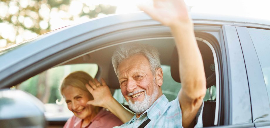 Ein Seniorenpaar sitzt in einem Auto und winkt.