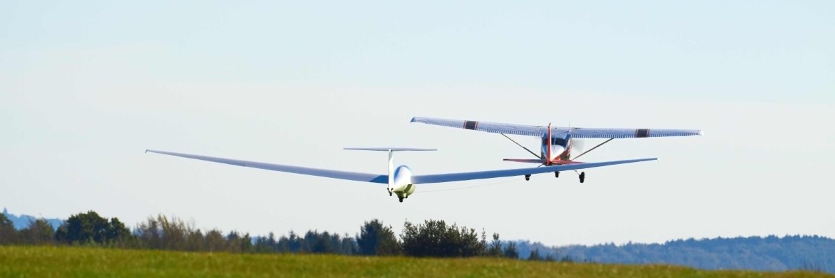 Zu sehen ist der Start eines Segelflugzeugs von einer grünen Wiese, welches von einem kleinen Flugzeug in die Luft gezogen wird. 
